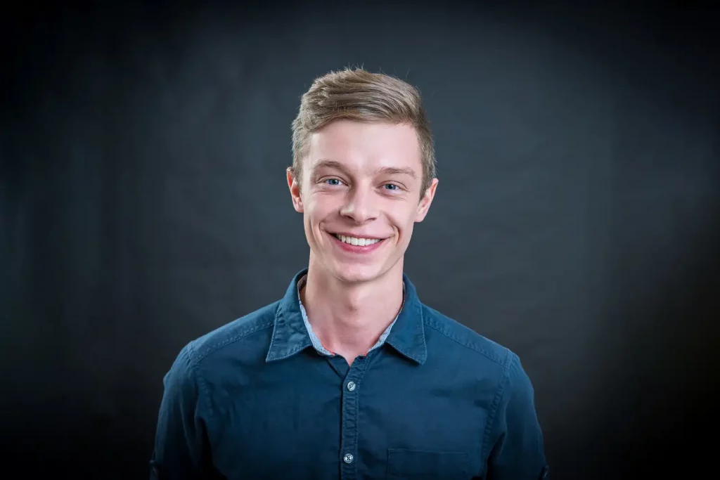 Ein junger Mann, Mitte 20, mit kurzen, dunkel blonden Haaren und einem breiten, freundlichen Lächeln wird hier abgebildet. Er trägt ein dunkelblaues Hemd mit offenem Kragen. Sein Ausdruck vermittelt Selbstbewusstsein, Kompetenz und eine positive Ausstrahlung. Der dunkle Hintergrund hebt ihn klar hervor.