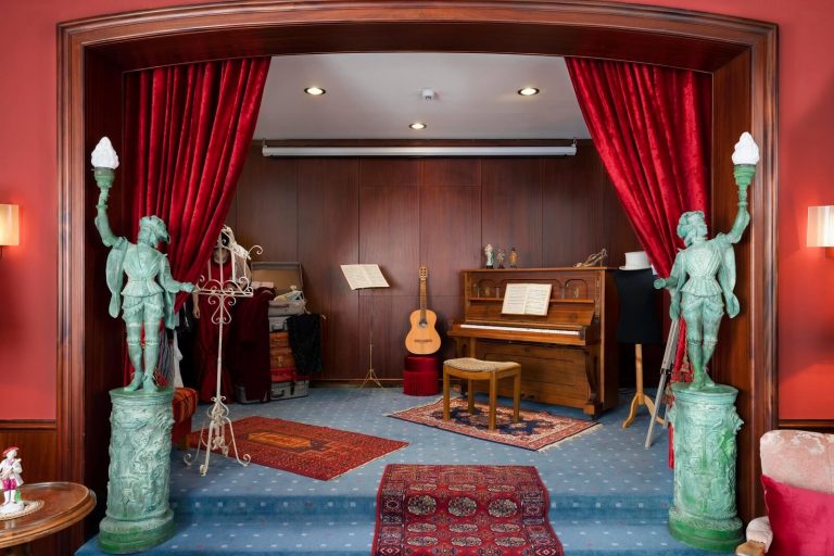 Bühnenbereich im Gemeinschaftsraum des Tante ALMA Hotels Mannheim mit roten Samtvorhängen, Klavier, Gitarre und nostalgischer Theaterkulisse